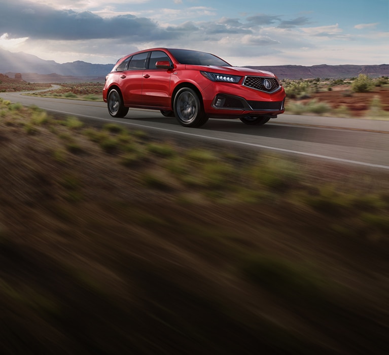 2019 Acura MDX A Spec in Performance Red Pearl front driving on a desert road under a cloudy sky (Premium Performance SUV). 2019 Acura MDX A Spec in Performance Red Pearl front driving on a desert road under a cloudy sky (Premium Performance SUV).